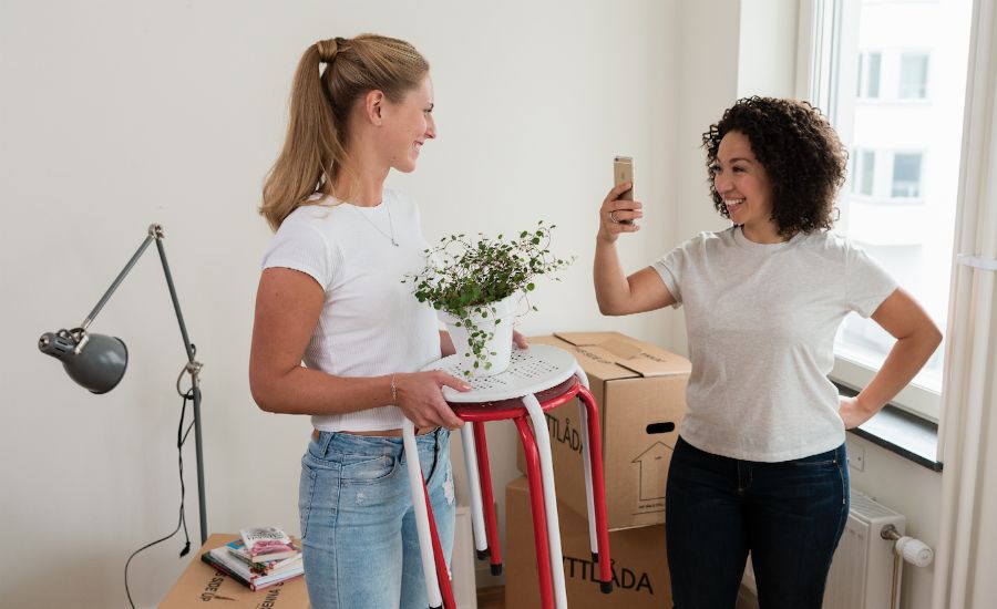 Två personer i ett rum med flyttlådor, en vit golvlampa och ett fönster; en person håller en pall med en krukväxt, medan den andra tar ett foto med en mobiltelefon