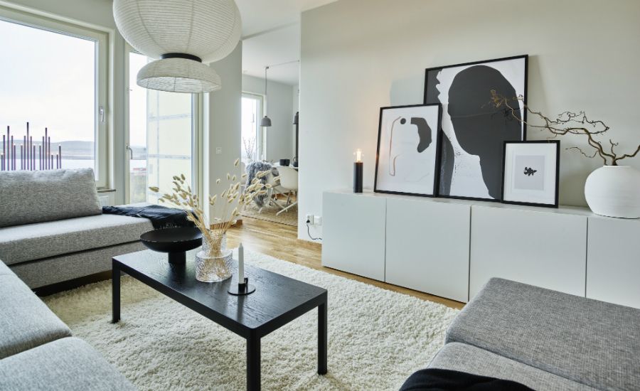 Modernt vardagsrum med grå soffor, svart soffbord, ljus matta och väggdekorationer. Stora fönster släpper in naturligt ljus