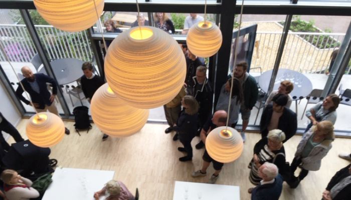 Vy ovanifrån av människor som samlats i ett modernt inomhusutrymme med stora fönster och flera hängande runda lampor med varmt ljus