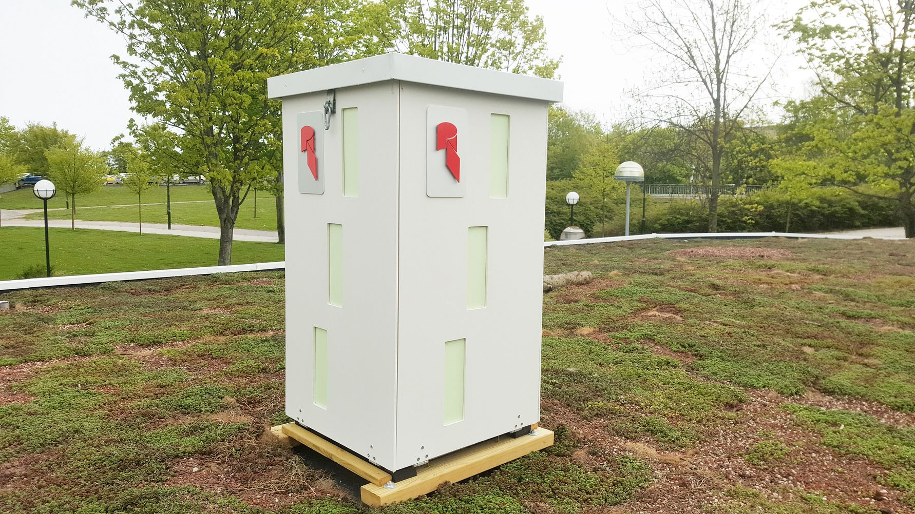 Vit fyrkantig låda med röda symboler på två sidor, placerad på ett grästak med träd och lampor i bakgrunden