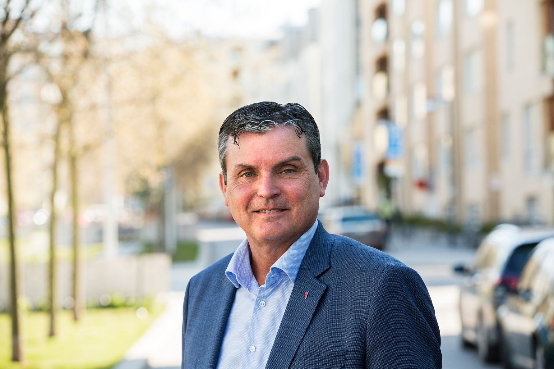 En man i blå kavaj och blå skjorta står utomhus på en stadsgata med träd och byggnader i bakgrunden