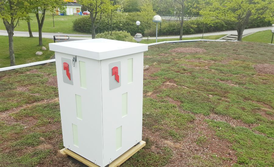 Vit rektangulär låda med röda symboler, placerad på ett grönt tak med vegetation och omgivande träd i bakgrunden