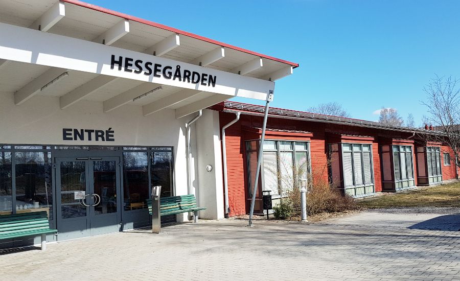 Entré till byggnaden Hessegården med glasdörrar, vita väggar och röda detaljer, omgiven av utomhusområde med bänkar och blå himmel