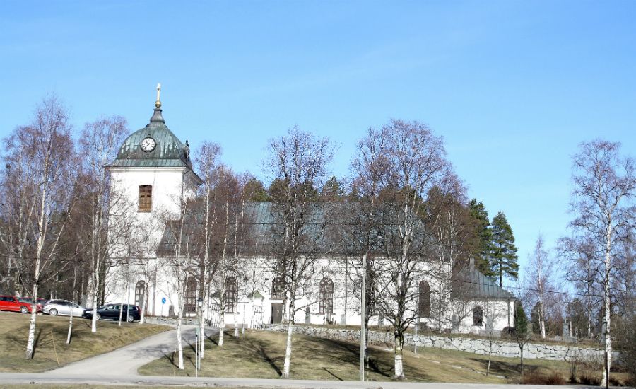 Vit kyrkobyggnad med koppartak och klocktorn, omgiven av nakna träd och en blå himmel i bakgrunden