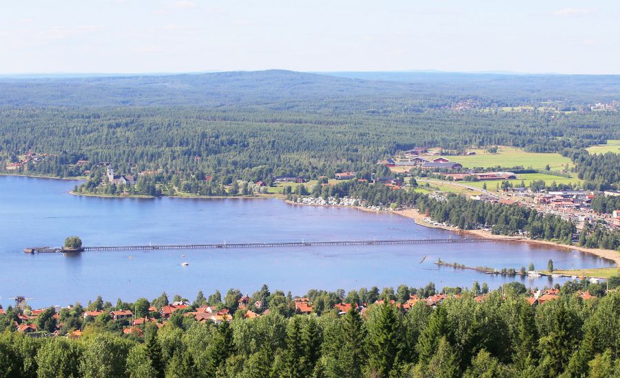 Utsikt över en sjö med en lång brygga, omgiven av skog och en liten stad i bakgrunden