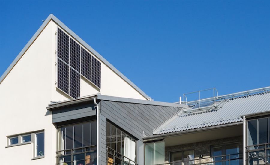 Vit byggnad med solpaneler monterade på taket, stora fönster och ett modernt arkitektoniskt designat tak mot en klarblå himmel