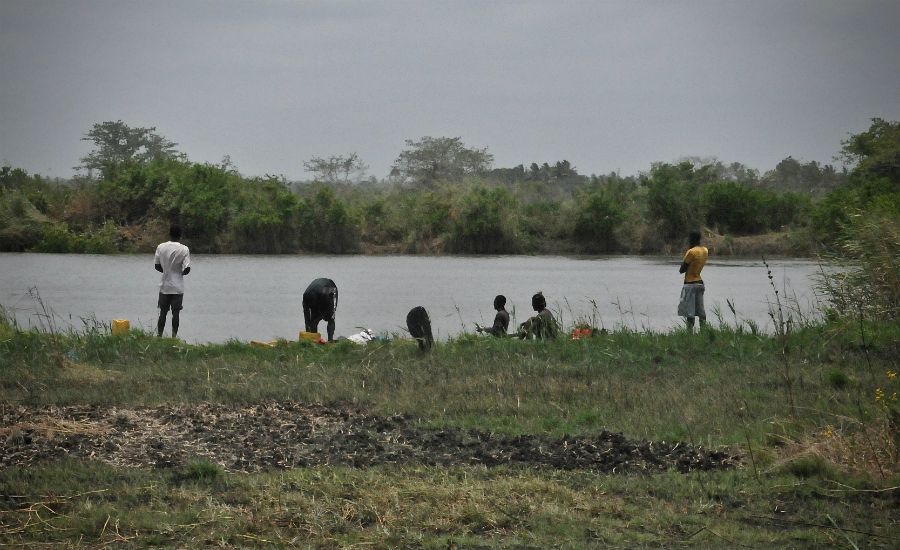 Personer vid en flodkant omgivna av grönska, några står och andra sitter eller böjer sig nära vattnet