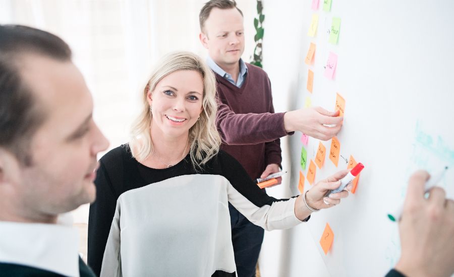 Personer står vid en whiteboard som är täckt med färgglada post-it-lappar och diskuterar idéer