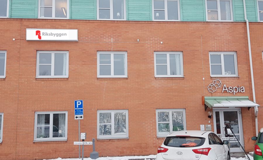 Byggnad med fasad av röd tegel, skyltar för Riksbyggen och Aspia, parkeringsskylt samt en vit bil parkerad framför. Snö täcker marken