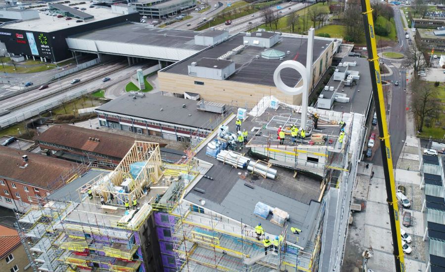 Byggarbetsplats med arbetare på ett flerfamiljshus under konstruktion, med byggställningar och material på taket. En kran syns bredvid byggnaden, omgiven av stadsmiljö