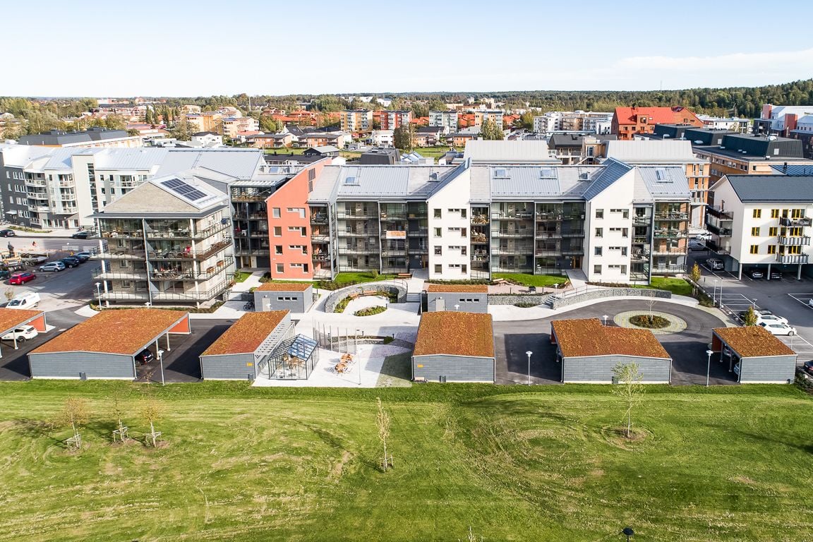 Modern bostadskomplex med flera byggnader, omgivna av grönområden och parkeringsplatser, med stadsvy i bakgrunden