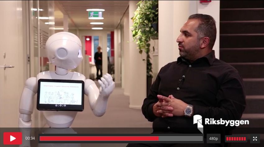Robot med en skärm står bredvid en sittande man i svart skjorta i en kontorskorridor. Riksbyggens logotyp syns i nedre högra hörnet