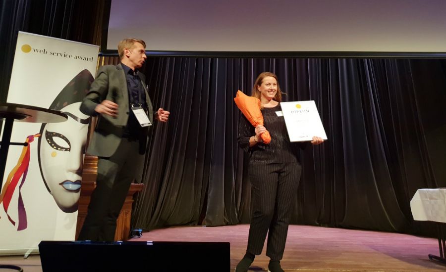 En person på en scen håller i en blombukett och ett diplom, bredvid en annan person. En banner med texten "web service award" syns i bakgrunden
