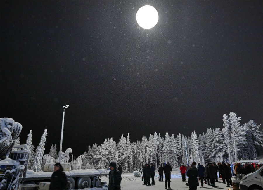 Snöklädd skog under natthimmel med en starkt lysande ballong ovanför och människor samlade på marken