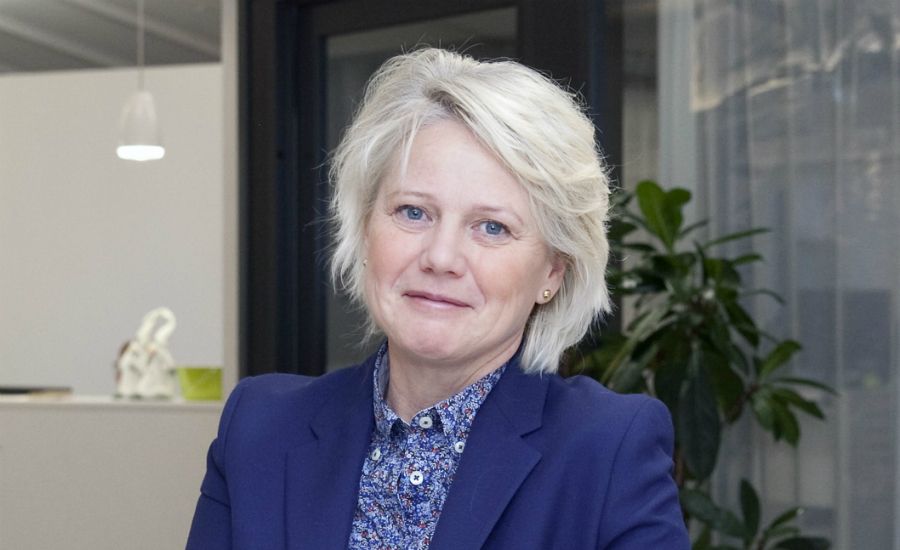 Porträtt av en person i blå kavaj och mönstrad skjorta, stående i en modern inomhusmiljö med växter och glasväggar