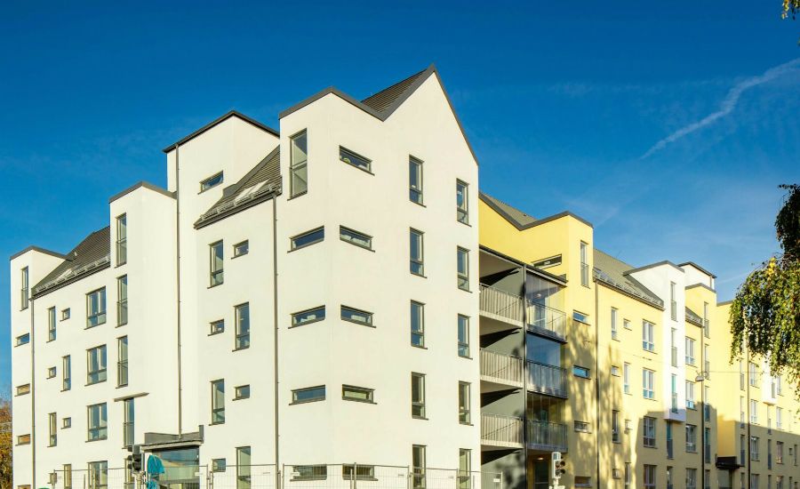 Vit modern flerfamiljsbyggnad med asymmetriska tak och stora fönster, omgiven av ett klart blått himmel och några träd