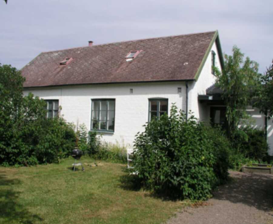 Vit enplanshus med sluttande tak, omgiven av grönska och buskar i en trädgård