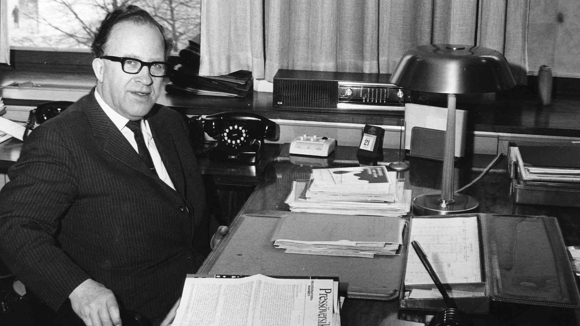 Man i kostym sitter vid ett skrivbord fyllt med papper och en skrivbordslampa