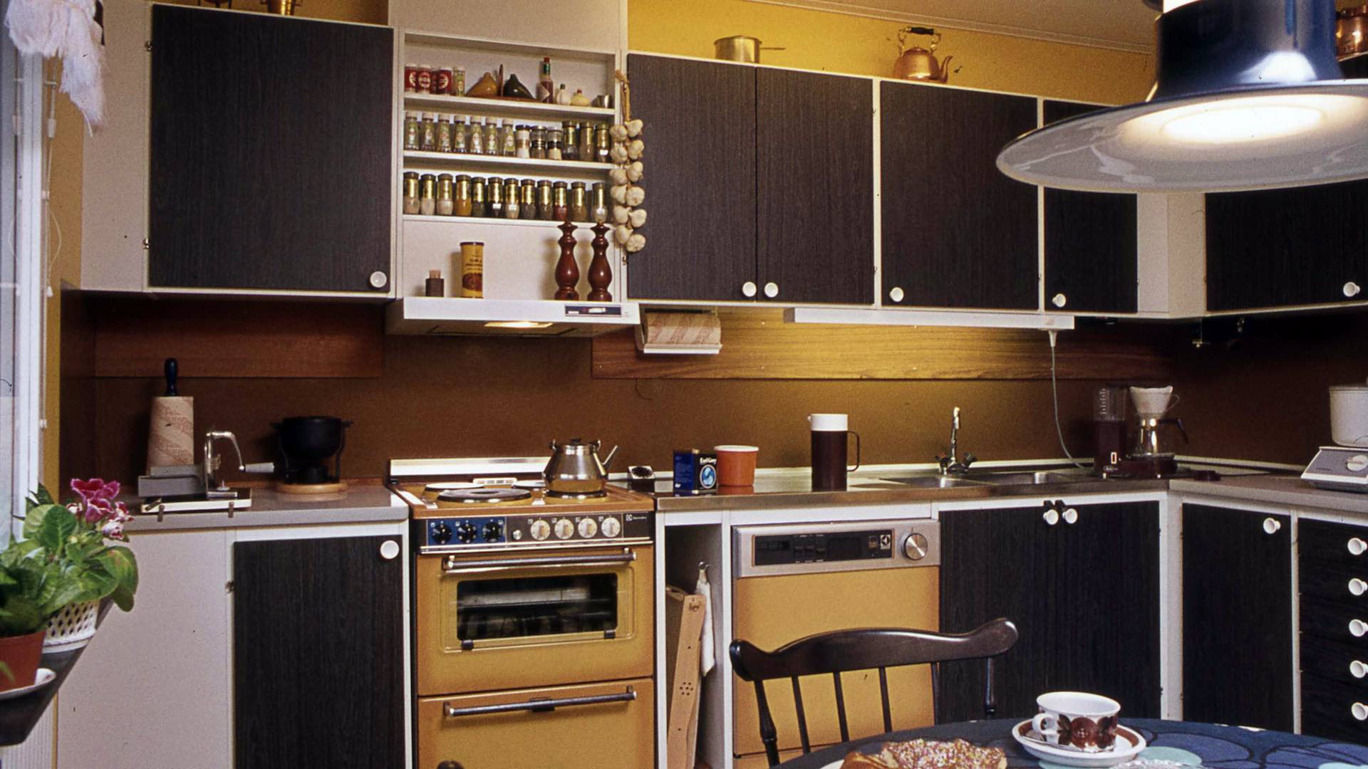 Kök i retrostil med bruna och vita skåp, en gul spis, kryddhylla, och en matplats med en svart stol och tallrikar