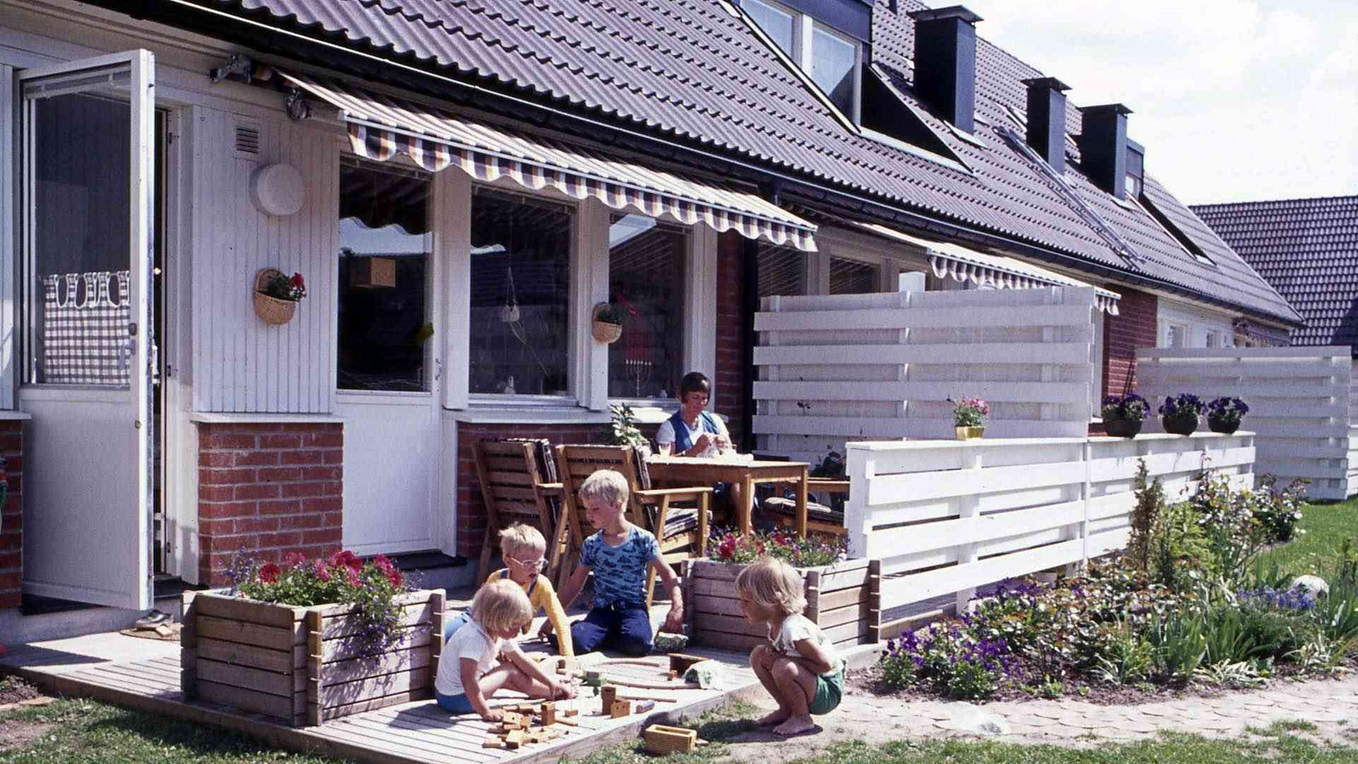 Radhus med tegelfasad och vita fönster. I förgrunden leker barn med klossar på altanen, en vuxen sitter vid ett bord i bakgrunden