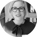 Kvinna med blont hår och glasögon, klädd i en mörk blus med rosett