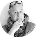 Porträtt av en man med glasögon på huvudet, iförd halsduk och blazer, med handen vid hakan