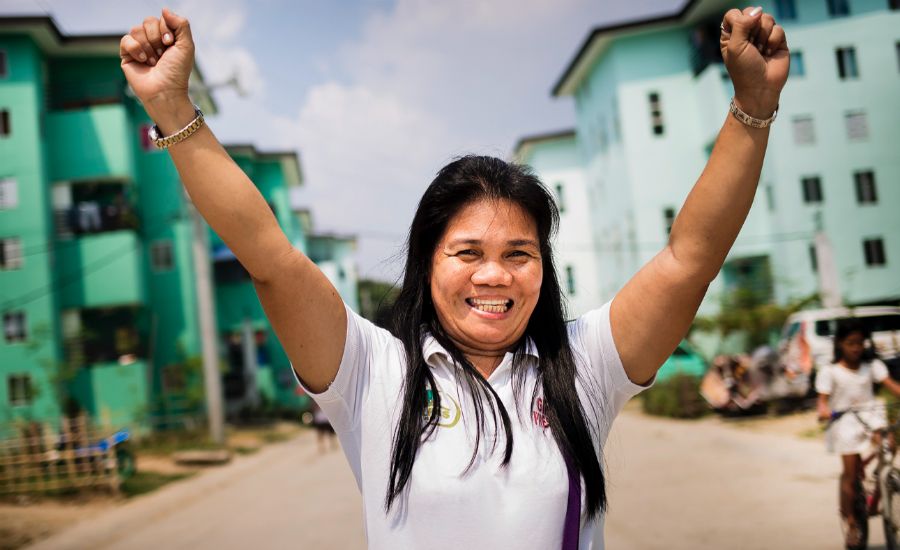 Person i vit kortärmad pikétröja som sträcker upp armarna i luften, med flerfamiljshus och blå himmel i bakgrunden