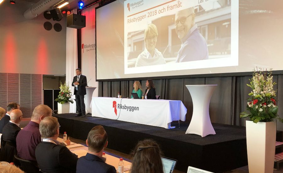 En konferenssal med en talare som står på scenen bredvid en projektorduk som visar en presentation. Ett panelbord med tre personer sitter framför scenen, omgiven av blommor och vit dekor