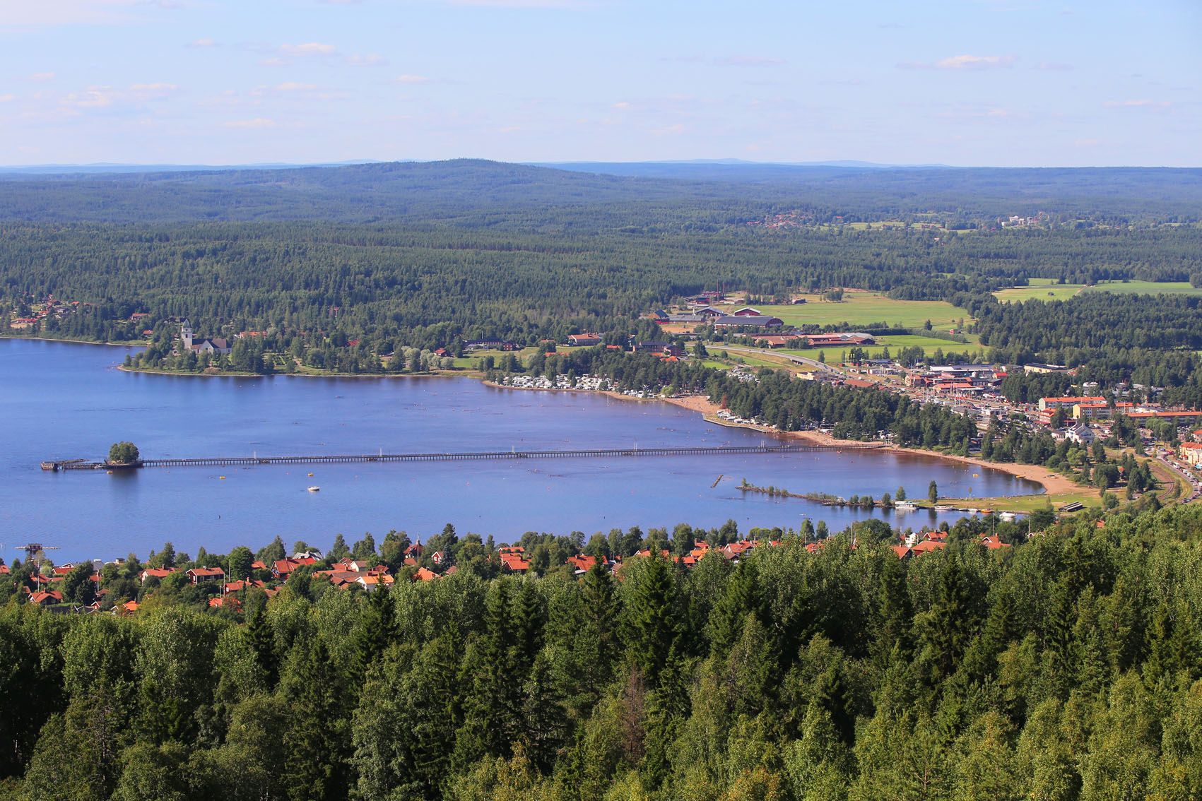 Utsikt över en sjö omgiven av skog och landsbygd, med hus och byggnader längs strandlinjen och en lång brygga som sträcker sig över vattnet