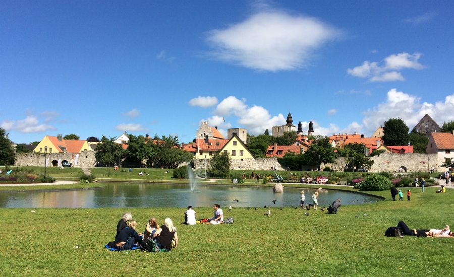 Gräsmatta med människor som sitter och njuter av en solig dag vid en damm, med stadsmurar och kyrktorn i Visby i bakgrunden