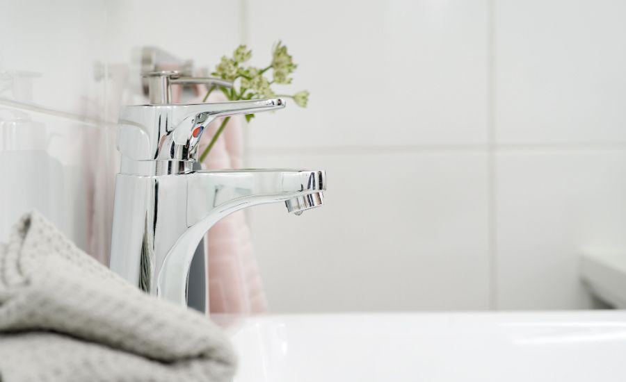 Kromad vattenkran på ett vitt handfat med en grå handduk och en vas med blommor i bakgrunden