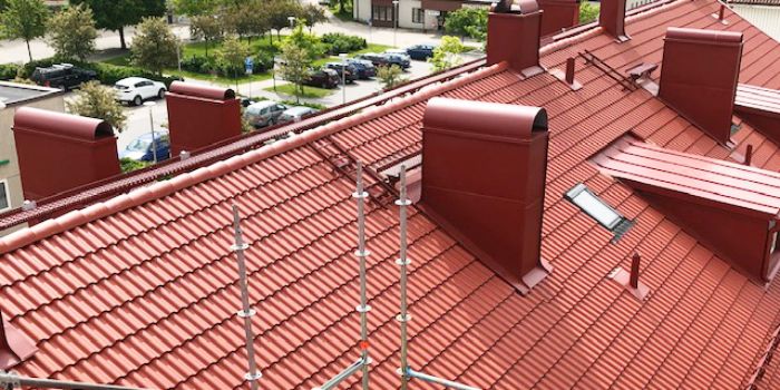 Rött tegeltak med flera skorstenar och ventilationsrör, sett från ovan. Byggnader och parkeringsplatser syns i bakgrunden