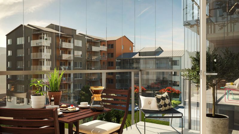 Balkong med glasväggar och utsikt över moderna lägenhetsbyggnader; möblerad med träbord, stolar, växter och en kaffebryggare