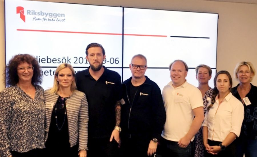 Gruppfoto framför en bildskärm som visar text från Riksbyggen, inklusive ett datum och logotyp. Personerna är klädda i olika typer av kontorskläder