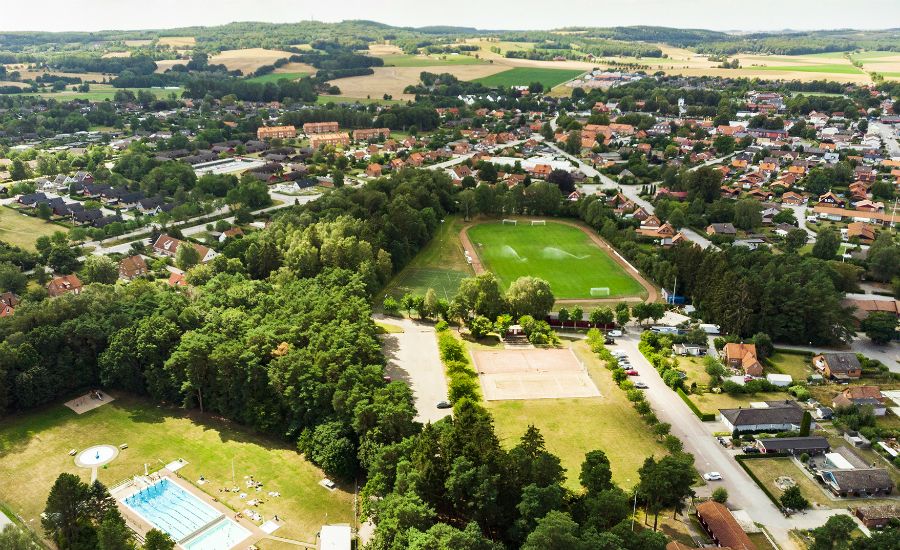Luftvy över en stad med en fotbollsplan, skogsområden, hus och en utomhuspool