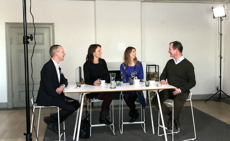 Fyra personer sitter vid ett bord i ett ljusrum med vita väggar och diskuterar. På bordet finns kaffekoppar, glas och anteckningar