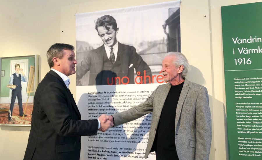 Två män skakar hand framför en utställning med text och bilder om Uno Åhrén