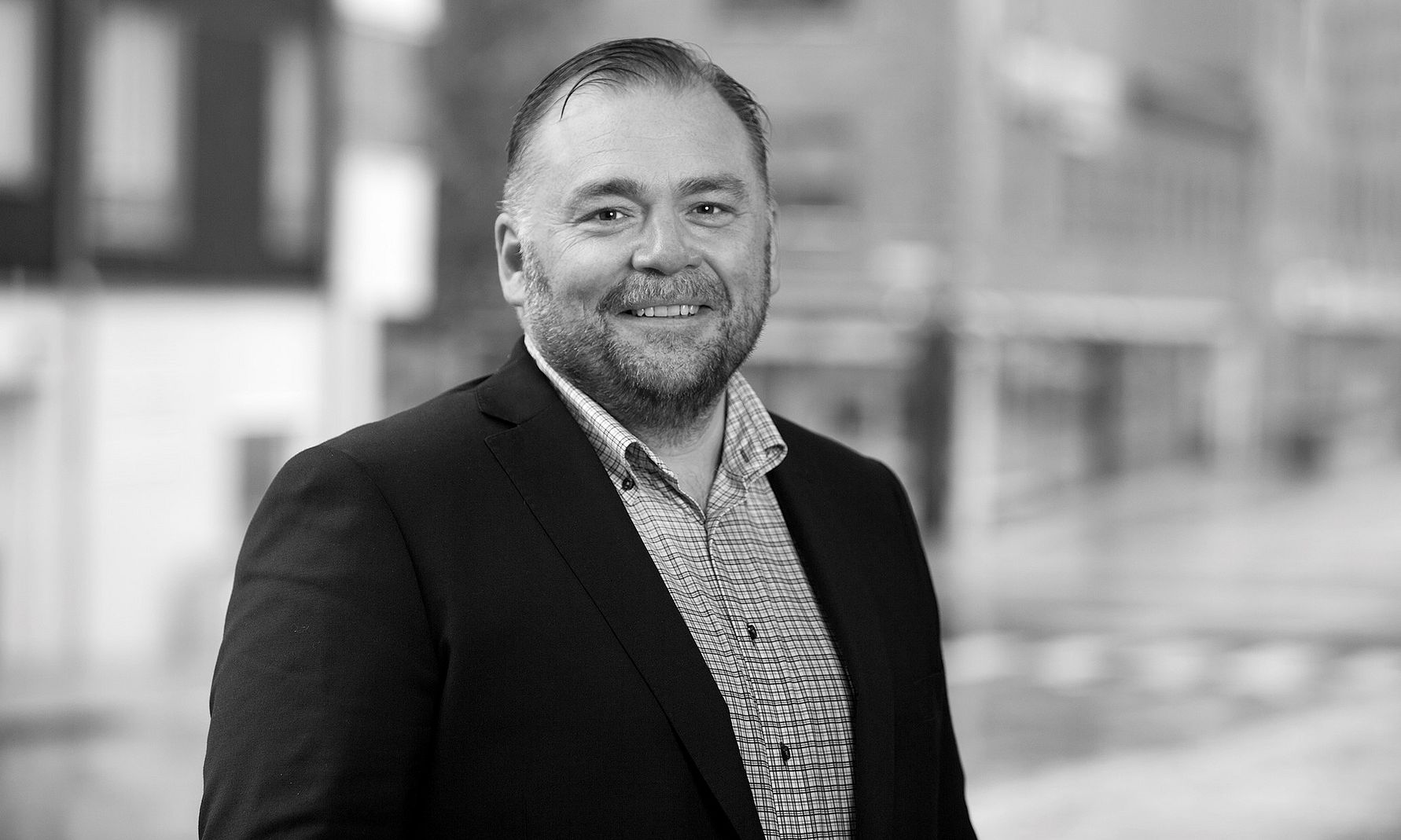 Man i kostym och rutig skjorta står utomhus med suddig stadsmiljö i bakgrunden