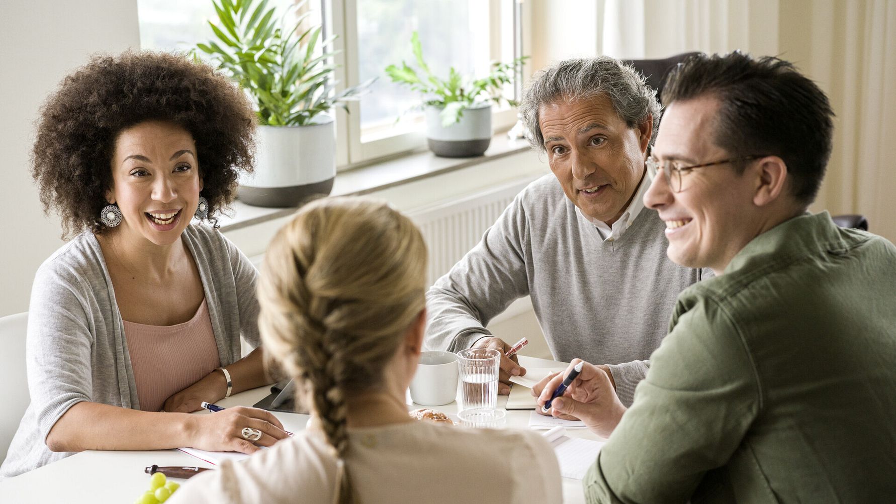 Fyra personer sitter runt ett bord i ett mötesrum med anteckningar, pennor och glas vatten framför sig. Gröna växter och ett fönster syns i bakgrunden