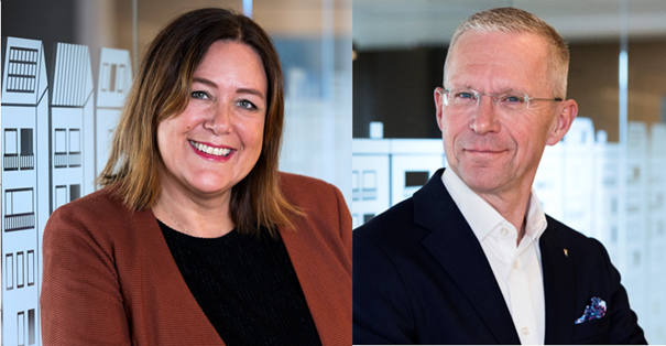 Två personer står bredvid varandra i en kontorsmiljö med glasväggar och vita dekorationer i bakgrunden. Den ena personen bär en brun kavaj och den andra en mörk kostym med vit skjorta