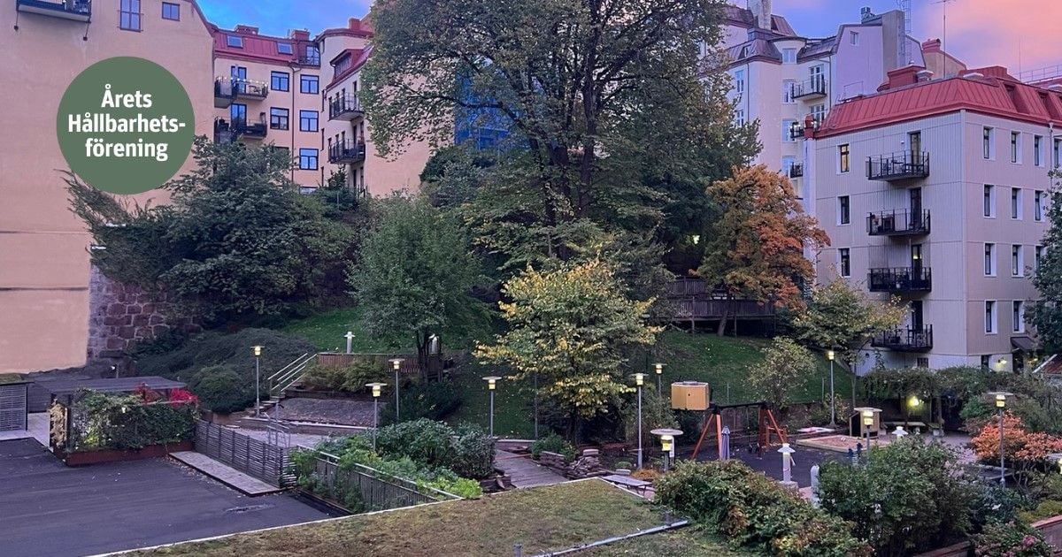 Flerfamiljshus med gröna uteområden och träd, omgivna av parkeringsplatser och belysning. En skylt med texten "Årets Hållbarhetsförening" syns till vänster