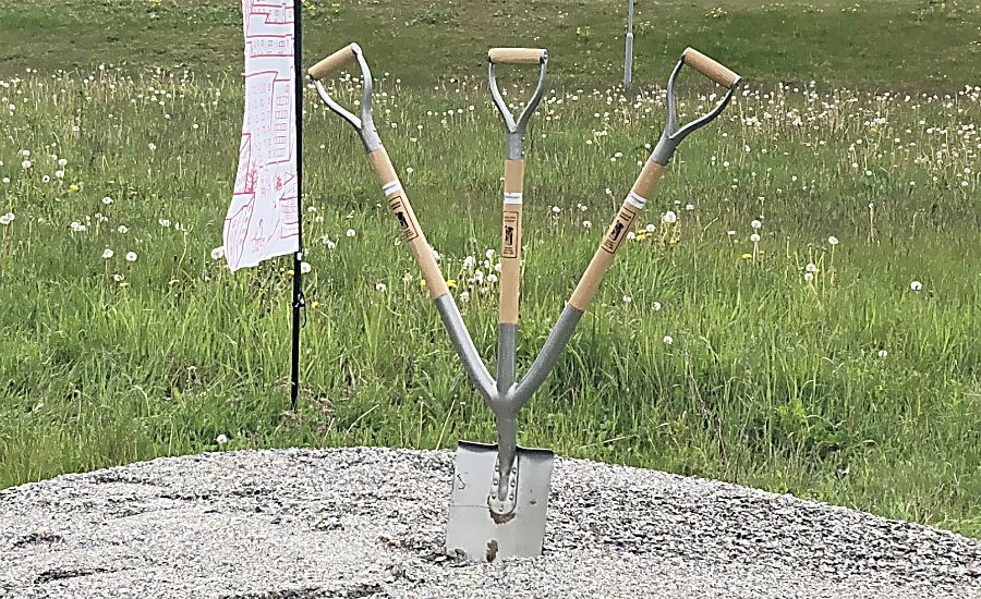 Tre spadar med trähandtag placerade i grus, omgivna av gräs och maskrosor. En skylt med text syns i bakgrunden