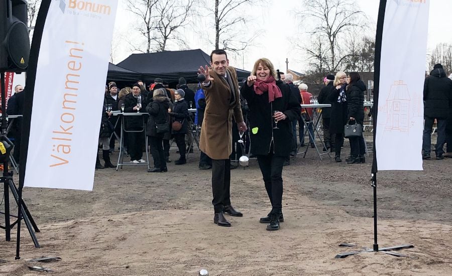 Två personer står framför en utomhuspublik mellan två vepor med texten "Välkommen!" på en marknads- eller evenemangsplats