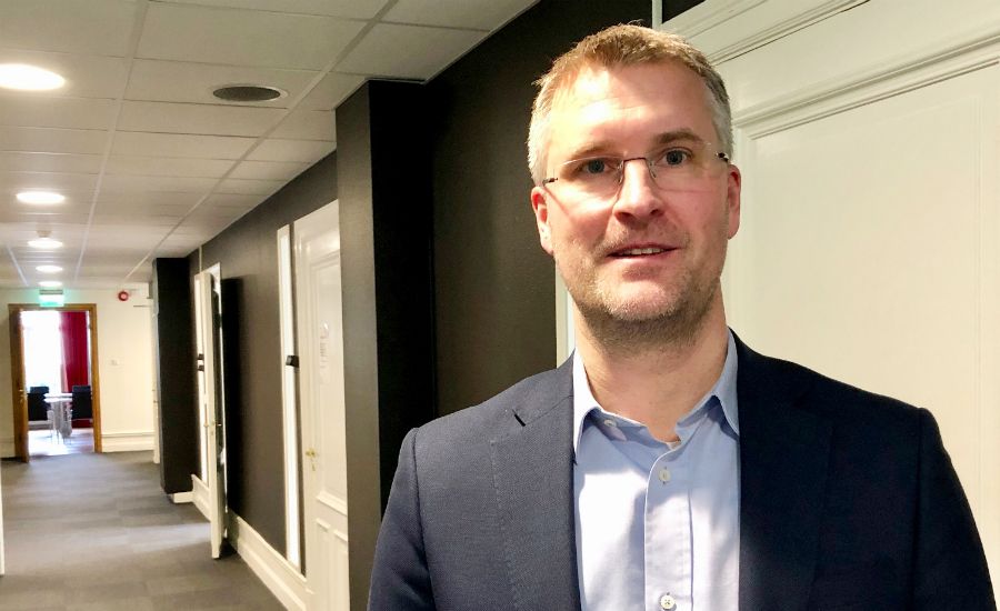 En man i mörk blazer och ljusblå skjorta står i en ljus korridor med vita dörrar och svarta väggar