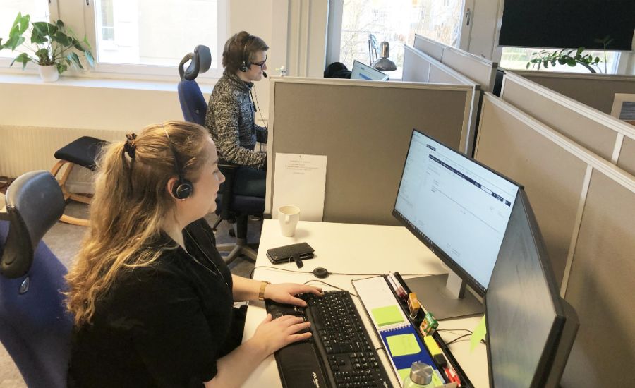 Personer arbetar vid skrivbord i ett kontorslandskap med datorer och hörlurar, omgivna av kontorsmöbler och växter