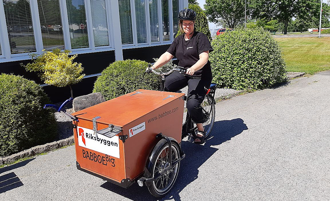 En person cyklar på en trehjulig lastcykel märkt med "Riksbyggen" och "BABBOE Big" framför en byggnad med glasfasad och omgivande gröna buskar