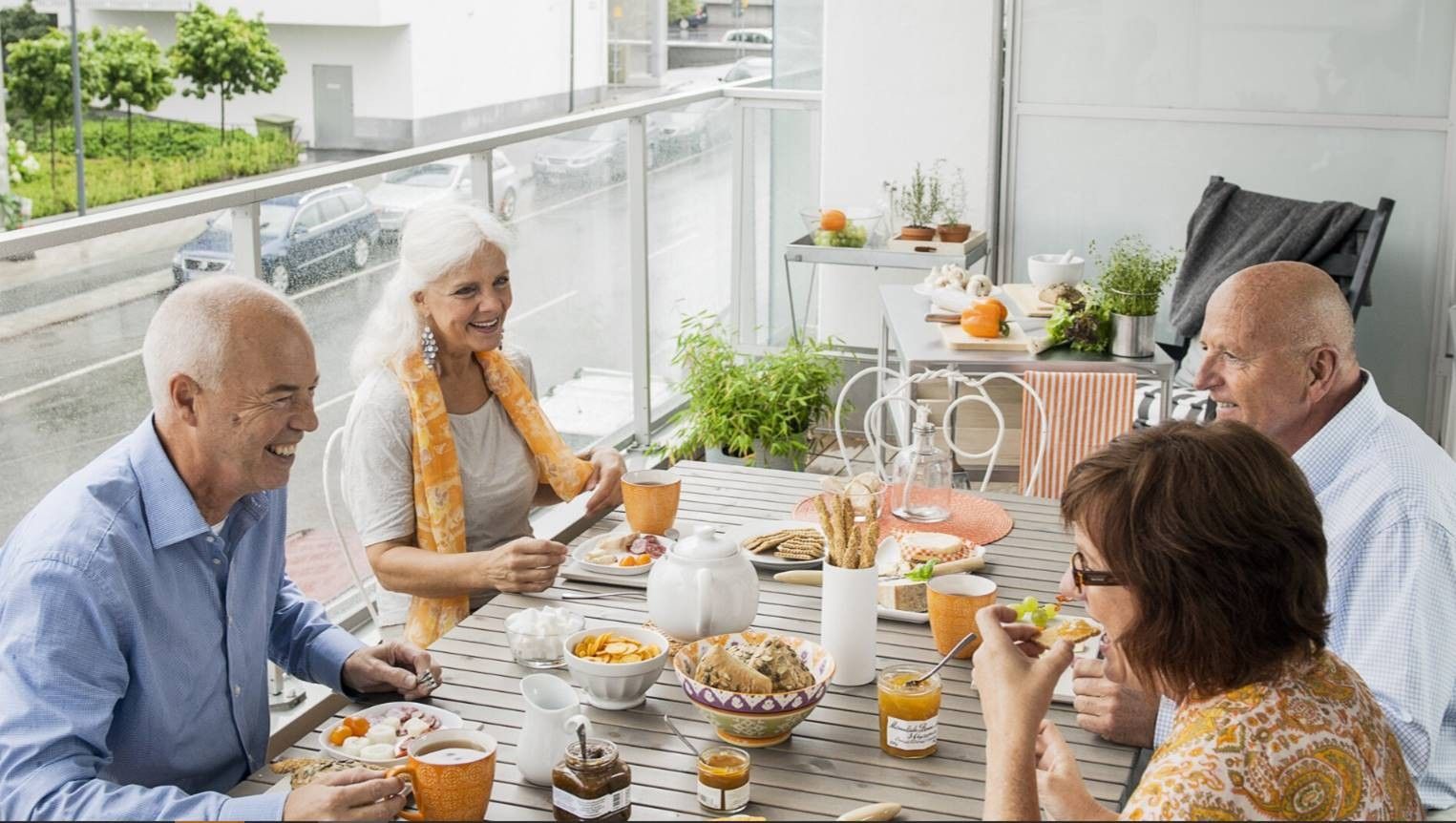 Fyra personer sitter vid ett dukat bord på en uteplats och äter en måltid. Bordet är dekorerat med orangefärgade muggar, tallrikar och olika maträtter