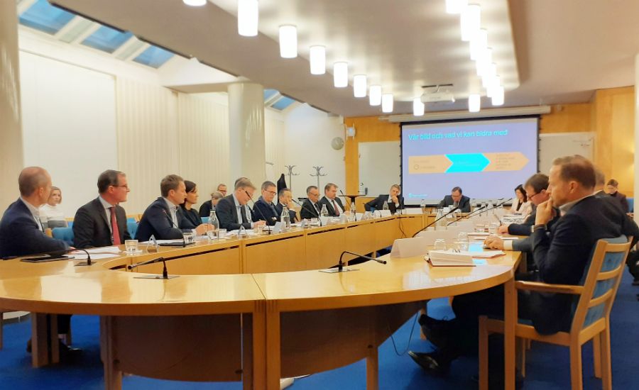 Möte i konferensrum med flera personer sittande runt ett stort runt bord, en presentation visas på en skärm i bakgrunden