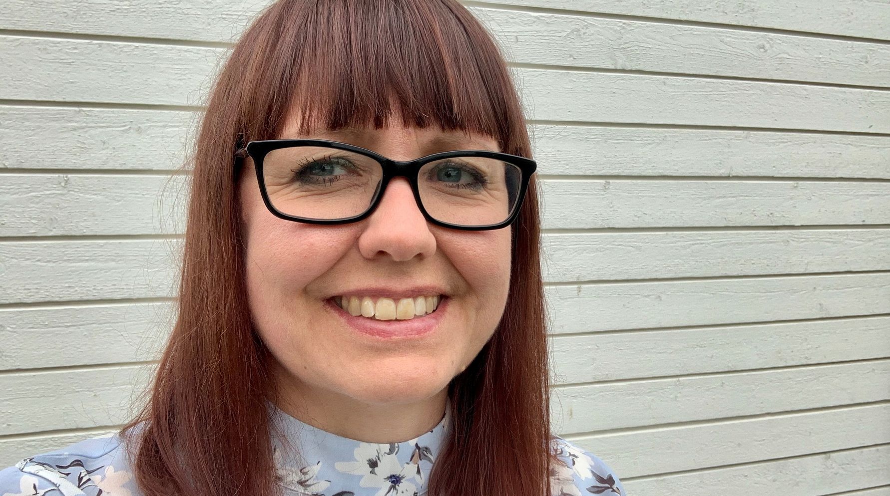 En person med långt rödbrunt hår och glasögon står framför en vit trävägg