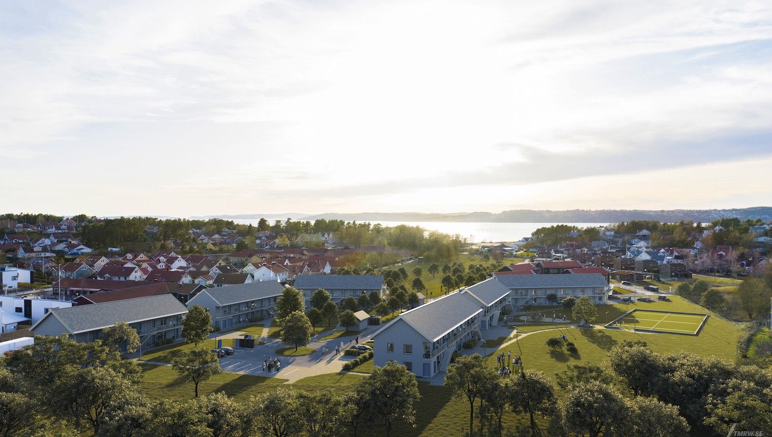 Flygbild av ett bostadsområde med flera byggnader, grönområden och träd, med havet och himlen i bakgrunden